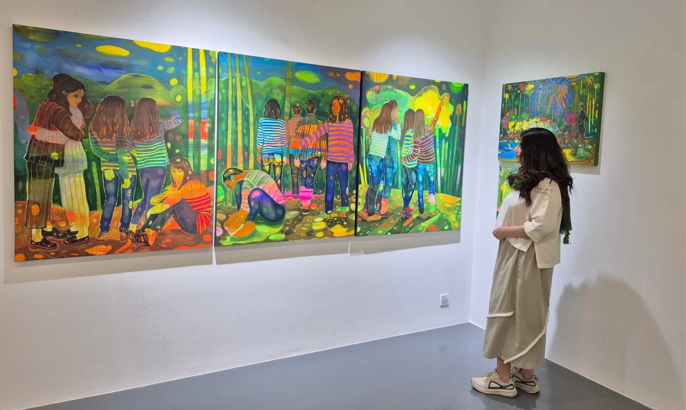 Mojgan looking at a bright triptych of young women in a forest, in a white gallery room